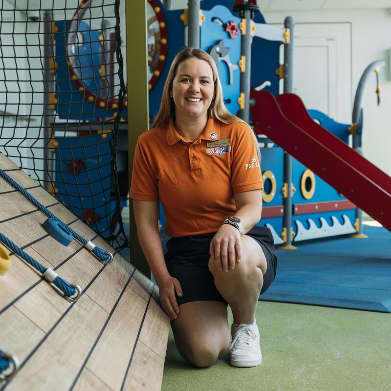 A woman in an orange polo shirt and navy shorts kneels and smiles in an indoor children's play area with climbing equipment and a blue and red slide in the background.