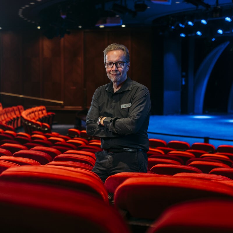 A man wearing glasses and a black shirt stands among rows of red theatre seats, smiling at the camera with an empty, dimly lit stage in the background.
