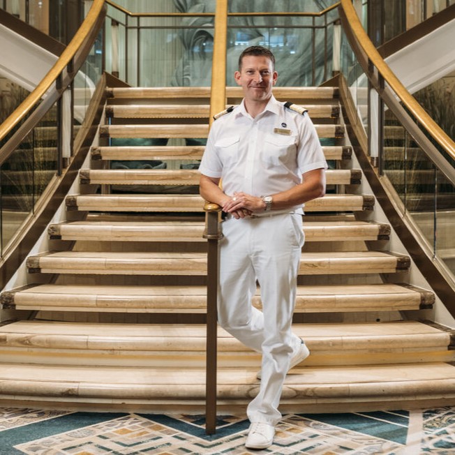 A man in a white naval uniform stands confidently at the base of an elegant, curved staircase with gold railings and patterned flooring in a luxurious interior setting.
