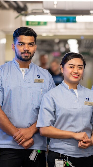 Two healthcare professionals wearing light blue uniforms and name badges stand side by side indoors, smiling at the camera. The background is softly blurred, suggesting a hospital or clinical setting.