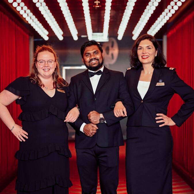 Three people dressed formally in black stand arm-in-arm, smiling, in a corridor with red curtains and bright ceiling lights, creating a glamorous, professional atmosphere.