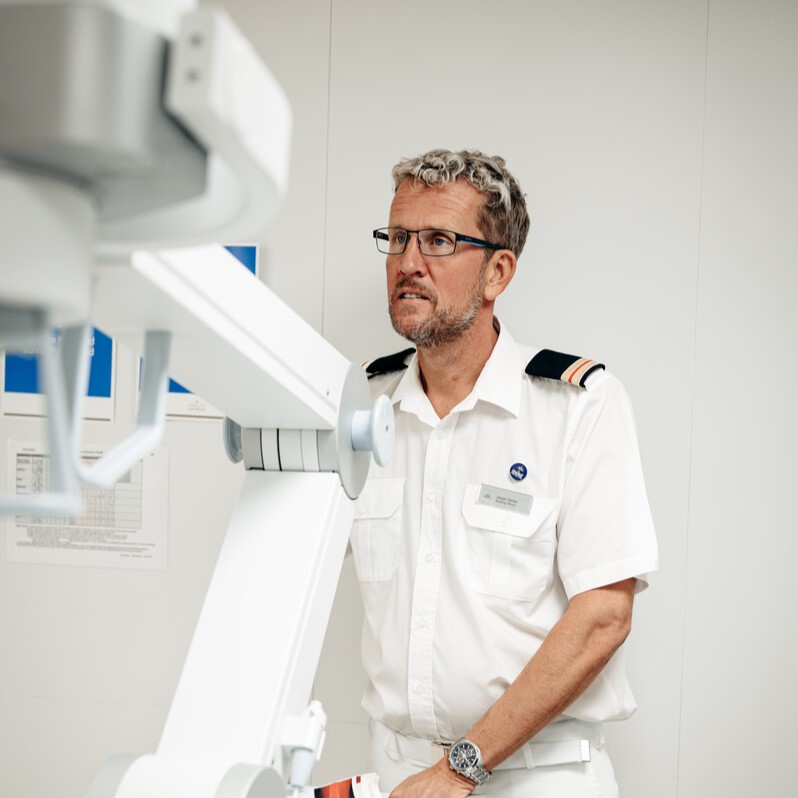 A man in a white uniform with shoulder stripes and a name badge stands next to medical equipment in a clinical setting, looking attentively at a screen.