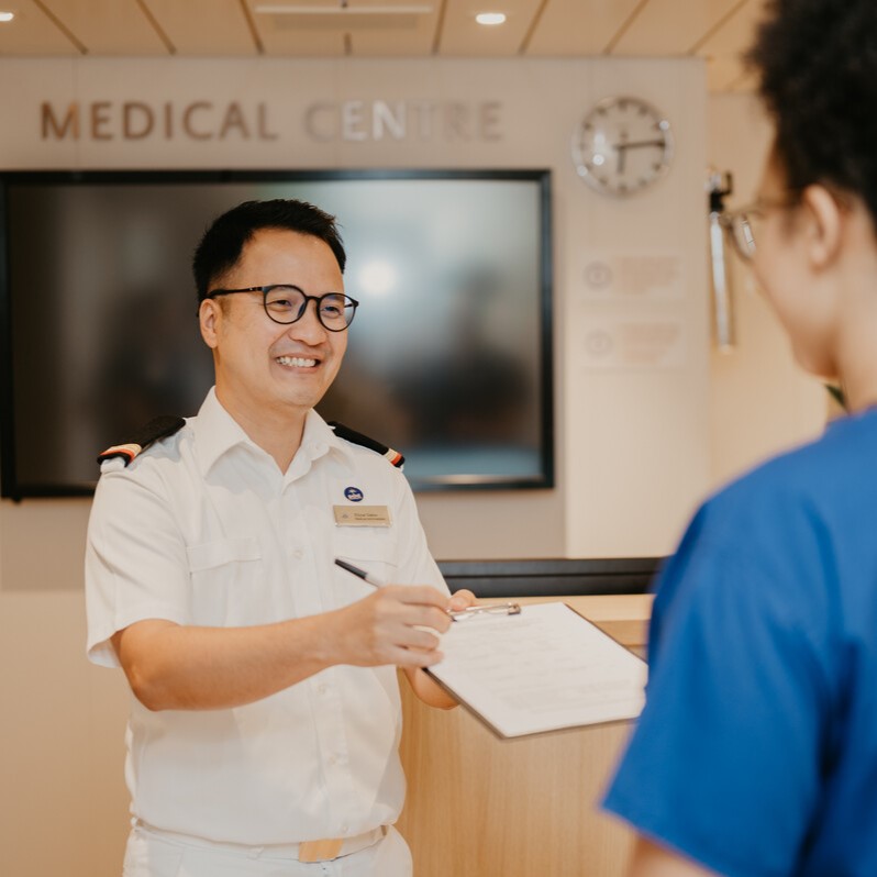 A smiling healthcare professional in a white uniform hands a clipboard to another person in blue scrubs at a medical centre reception desk. A clock and a large screen are visible in the background.
