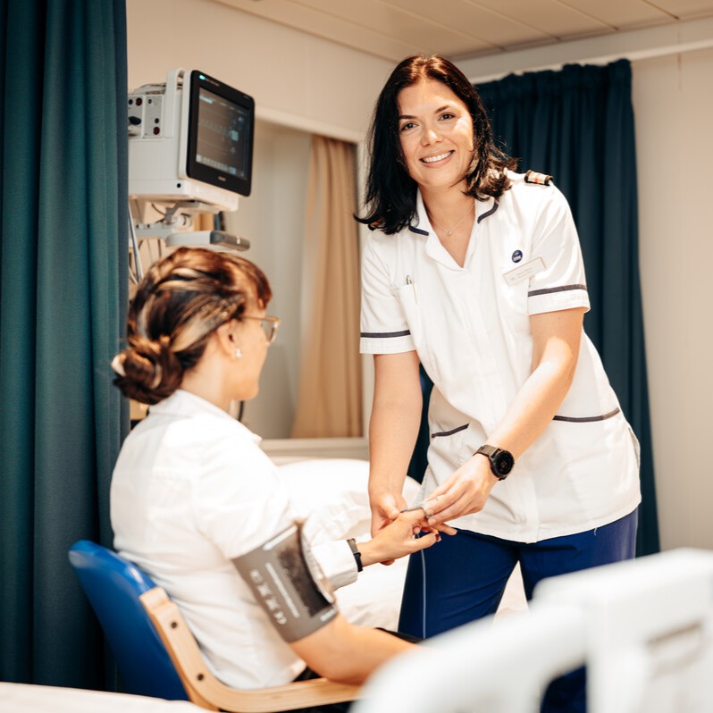 A nurse wearing a white uniform smiles whilst checking the blood pressure of a seated patient, also in uniform, in a medical setting with monitoring equipment in the background.