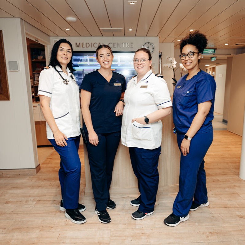 Four healthcare professionals, three in blue uniforms and one in white, stand smiling together in a medical centre with wood flooring and a sign reading Medical Centre in the background.