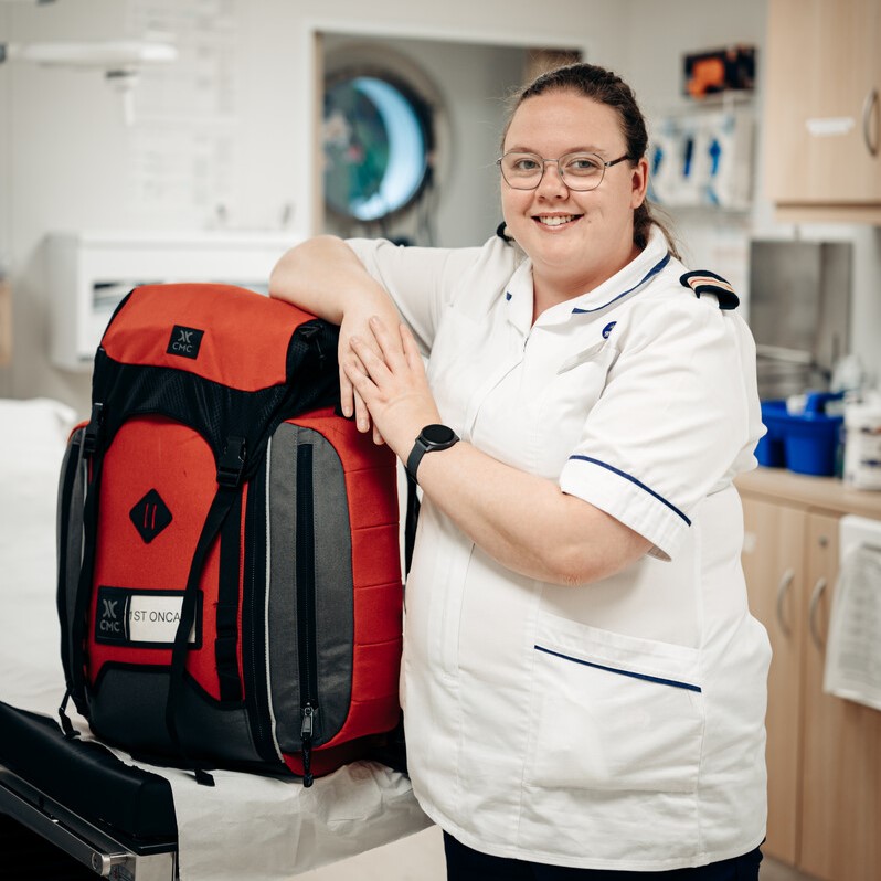 A smiling nurse in a white uniform stands in a medical room, resting her arm on a large red and black emergency bag placed on a hospital bed.