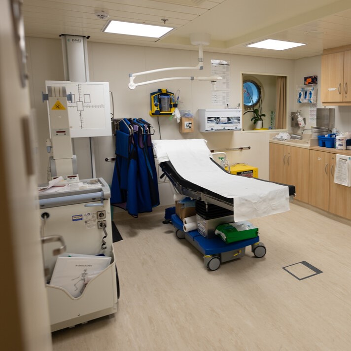 A clean, modern medical examination room with an examination couch in the centre, medical equipment, cupboards, and supplies along the walls, and a round window letting in natural light.