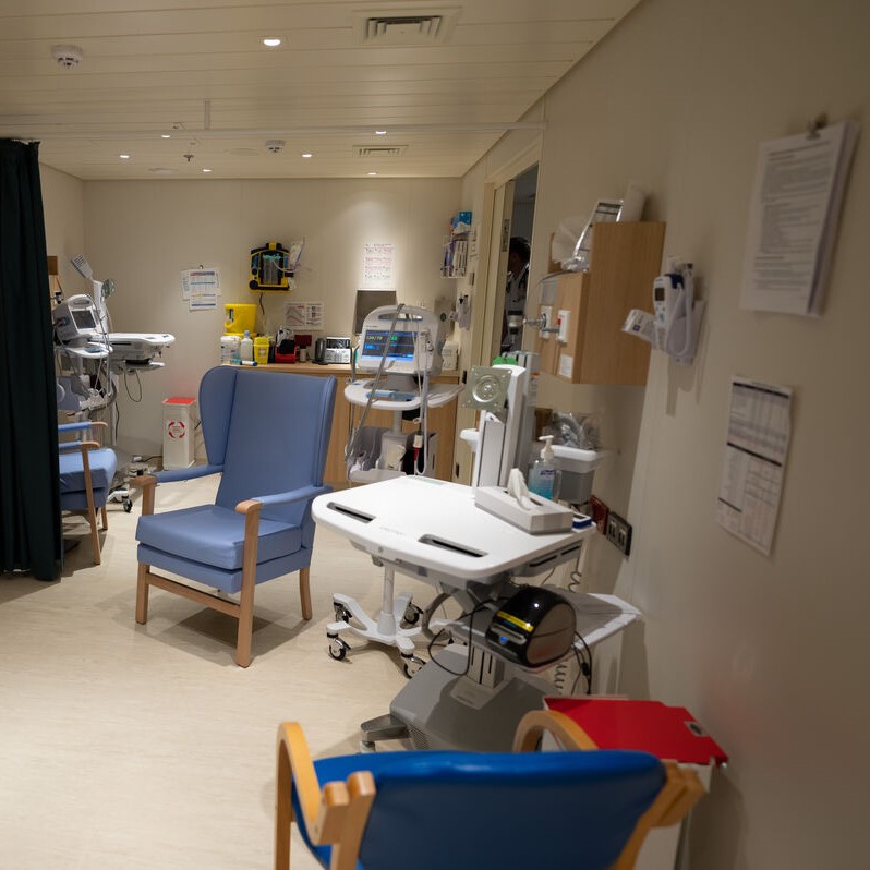 A hospital ward with medical equipment, blue chairs, workstations on wheels, monitors, and supplies on walls. Lighting is bright, and the room appears clean and organised, designed for patient care.