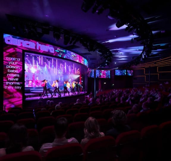 A vibrant stage performance called Dance Hero takes place in a theatre with colourful lights and dancers, while an audience watches from red seats. Song lyrics are displayed on screens above the stage.