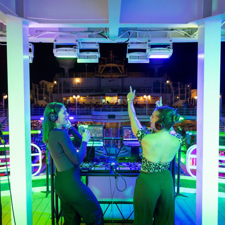 Two women DJ at a colourful, nighttime outdoor event on a ship’s deck. One holds a microphone, the other raises her arms, both wearing headphones. Bright lights and people are visible in the background.