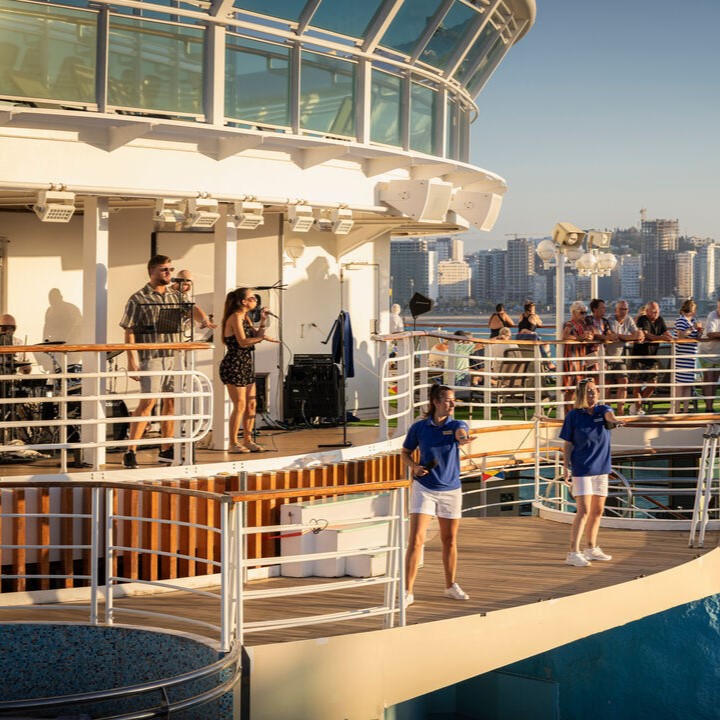 A band performs on the deck of a cruise ship as guests watch. Two staff members in blue shirts and white shorts dance in the foreground. A city skyline is visible in the background under a clear sky.