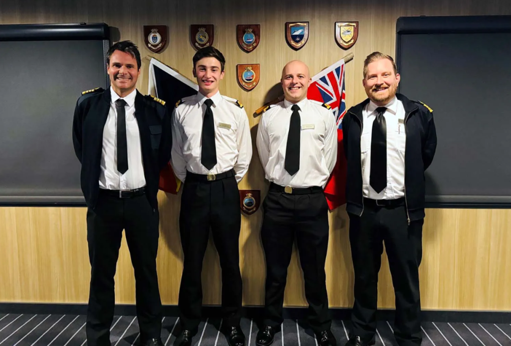Four smiling men in pilot uniforms stand side by side in front of a wood-panelled wall with shields and two flags, posing for a group photo indoors.
