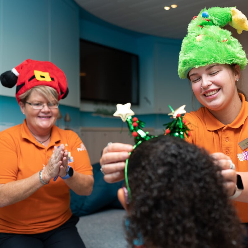 Two women in orange shirts, one wearing a Santa hat and the other a decorated green holiday hat, help a child put on festive headbands with star decorations. Everyone appears to be smiling and enjoying the activity.