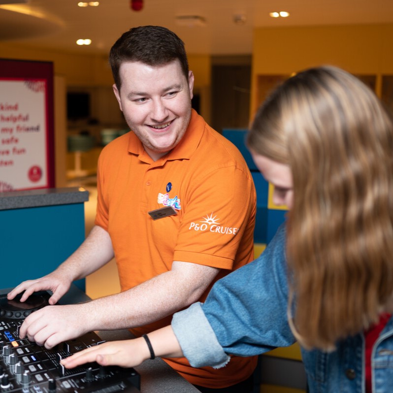 A smiling man in an orange P&O Cruises shirt helps a young woman operate a DJ mixer indoors, guiding her as she adjusts the controls.