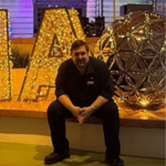 A man in dark clothing sits on a ledge in front of large illuminated letters and a decorative spherical structure, with colourful lighting in the background.