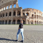A person poses in front of the Colosseum in Rome, Italy, on a sunny day. The ancient stone amphitheatre stands prominently in the background under a clear blue sky.