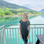 A woman stands on a balcony overlooking a calm lake with green hills and scattered houses in the background under a partly cloudy sky.