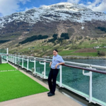 A person stands on the deck of a ship beside a river or fjord, with snow-capped mountains and partly cloudy skies in the background. Green artificial turf covers part of the deck.
