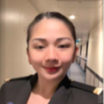 A person in a dark uniform with shoulder epaulettes is smiling at the camera whilst standing in a brightly lit corridor.