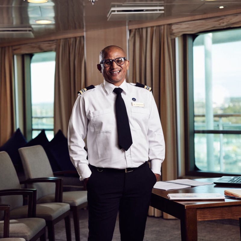 A man in a cruise ship officer’s uniform stands smiling beside a desk in a bright, modern room with large windows, beige curtains, and several chairs. Papers and a pen are on the desk.