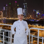 A chef in a white uniform and tall hat stands on a deck with a city skyline lit up at night in the background.