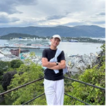A person in a black shirt and white trousers stands smiling in front of a scenic view with greenery, a body of water, cruise ships, and mountains in the background under a cloudy sky.
