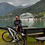 A man sits on a wooden bench beside a white bicycle near a calm lake with a large cruise ship and mountains in the background under a cloudy sky.