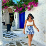 A woman in a white crop top and blue-patterned skirt walks on a stone path in a white-walled alley with vibrant pink flowers and a blue door, likely in a Mediterranean setting.