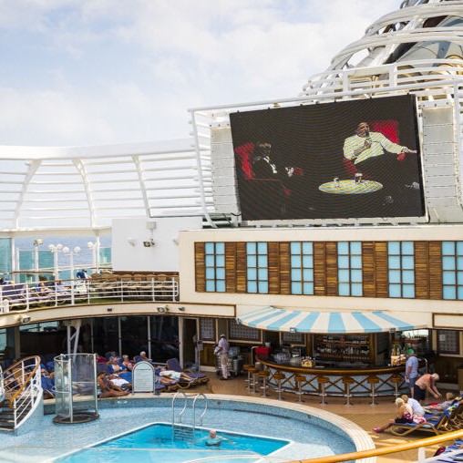 Passengers relax around a pool on a cruise ship while a film plays on a large outdoor screen above the deck. Some people lounge in chairs, and others walk nearby under a partly cloudy sky.