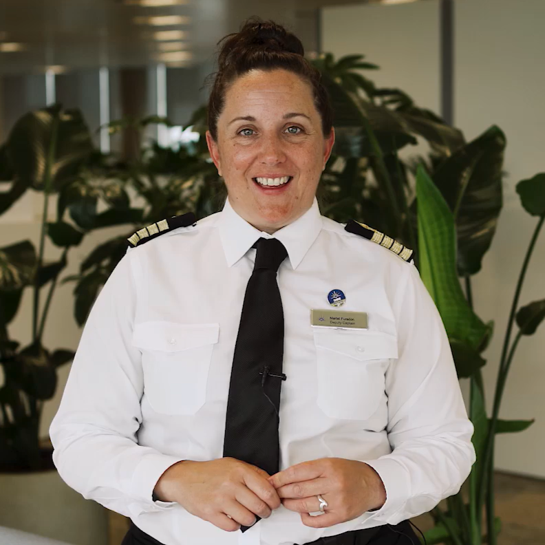 A smiling woman in a pilot’s uniform with shoulder epaulettes and a black tie sits indoors, in front of large green plants. She has brown hair tied up, a name badge, wedding ring, and embodies the professionalism of P&O Cruises deck careers.
