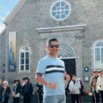A man wearing sunglasses and a light blue shirt stands in front of a stone building with arched windows. Several people are gathered in the background on a sunny day.