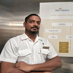 A man in a white uniform stands with folded arms in front of a P&O Cruises leadership organisation chart, looking at the camera.