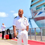 A man in a white naval or cruise ship uniform stands on a quay near a large cruise ship; he holds a mobile and is surrounded by luggage and other people under a bright, clear sky.