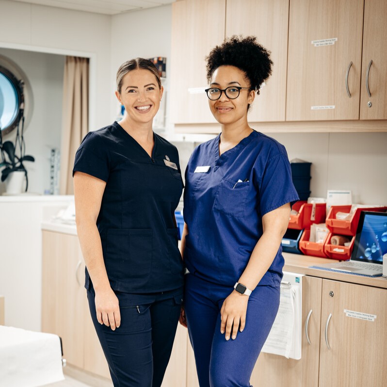 Two healthcare professionals in navy scrubs stand and smile in a medical office with wooden cupboards, medical supplies, and a round window in the background.