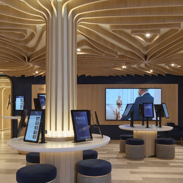 Modern interior space with wooden ceiling design, digital kiosks on round tables, cushioned stools, a central illuminated column, and a large wall display screen showing people in conversation.