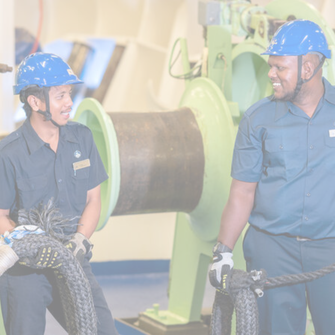 Two workers in blue uniforms and safety helmets stand near industrial machinery, holding thick ropes and smiling at each other—capturing the teamwork found in P&O Cruises deck careers, all within a bright indoor setting.
