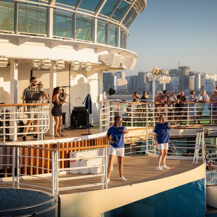 A live band performs on a cruise ship deck as two staff members interact with guests below. Passengers watch from upper and lower levels, with a city skyline visible in the background under a clear sky.