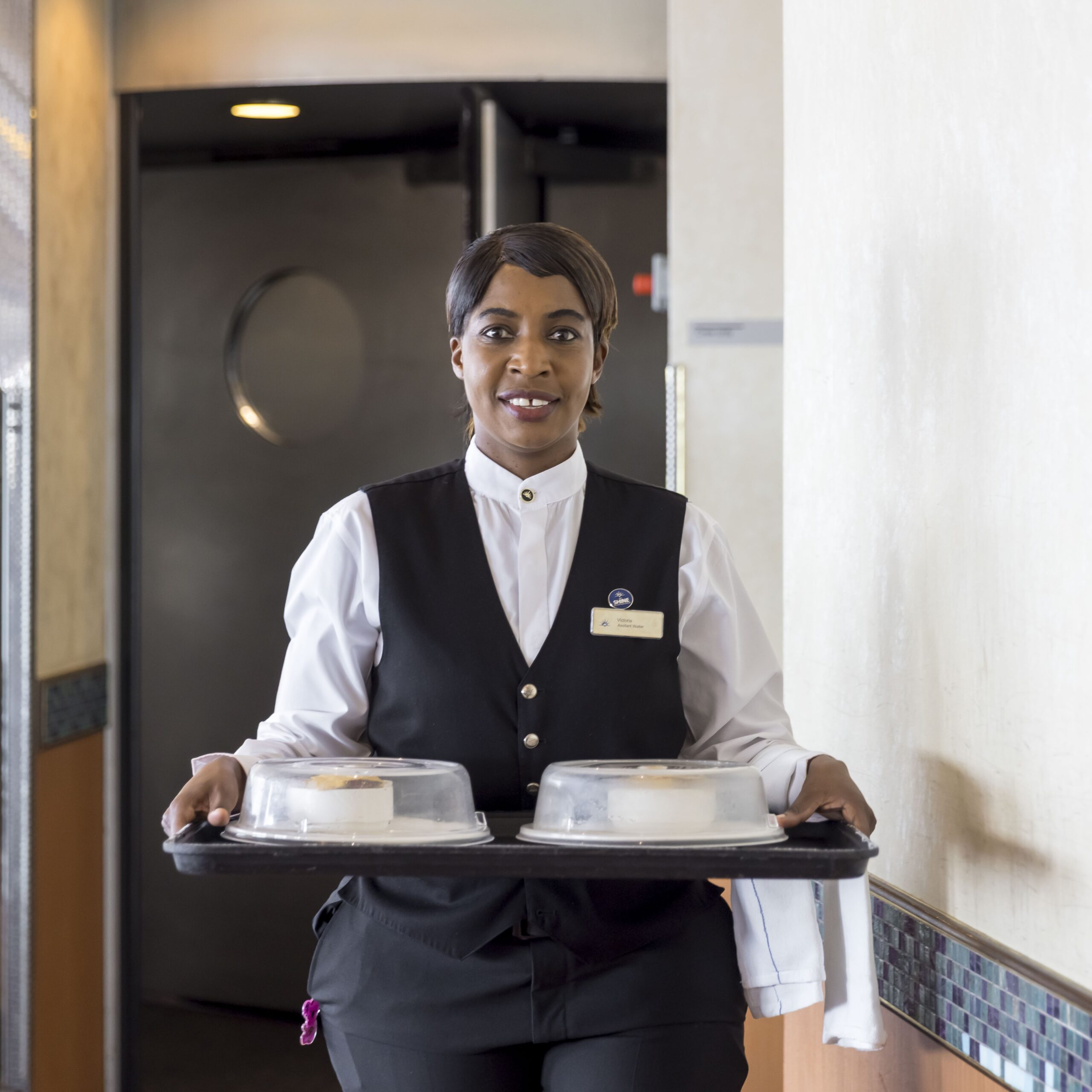 A restaurant waiter in a black waistcoat and white shirt carries a tray with two covered dishes, smiling as she walks through a dining area—showcasing the service excellence found in cruise ship restaurant jobs.