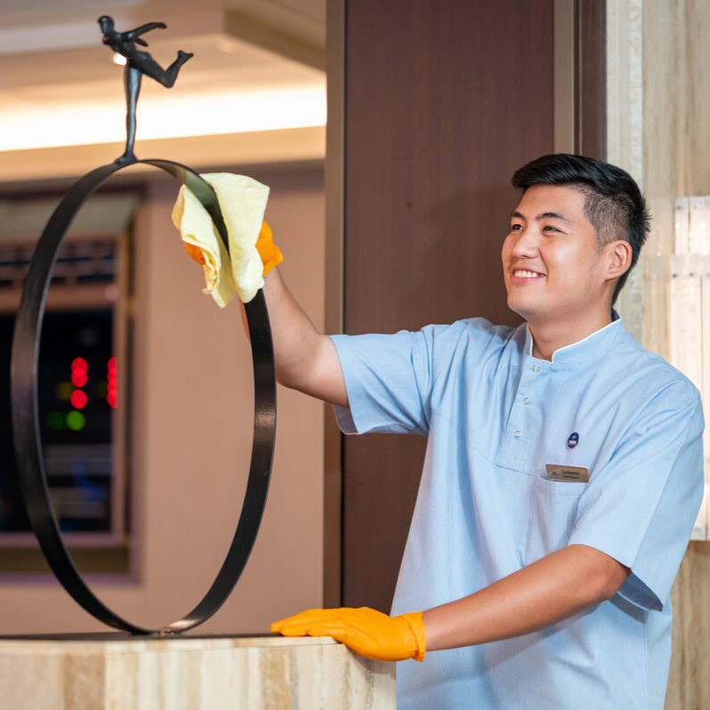 A smiling hotel staff member wearing orange gloves and a light blue uniform cleans a decorative metal sculpture with a yellow cloth in a well-lit interior.