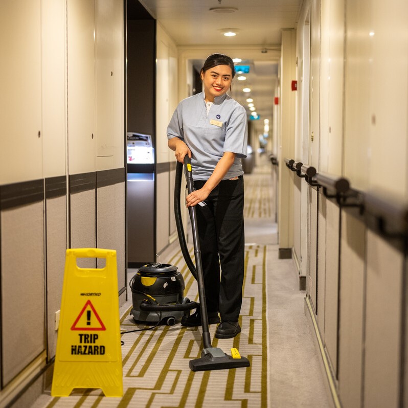 A smiling housekeeper hoovers a carpeted corridor. A yellow Trip Hazard warning sign is placed nearby. The corridor has cream walls, handrails, and doors on both sides.
