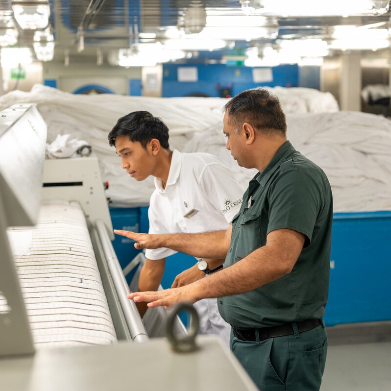 Two men, one in a white uniform and one in a green uniform, inspect and discuss a large industrial laundry machine in a brightly lit laundry facility with piles of folded linen in the background.