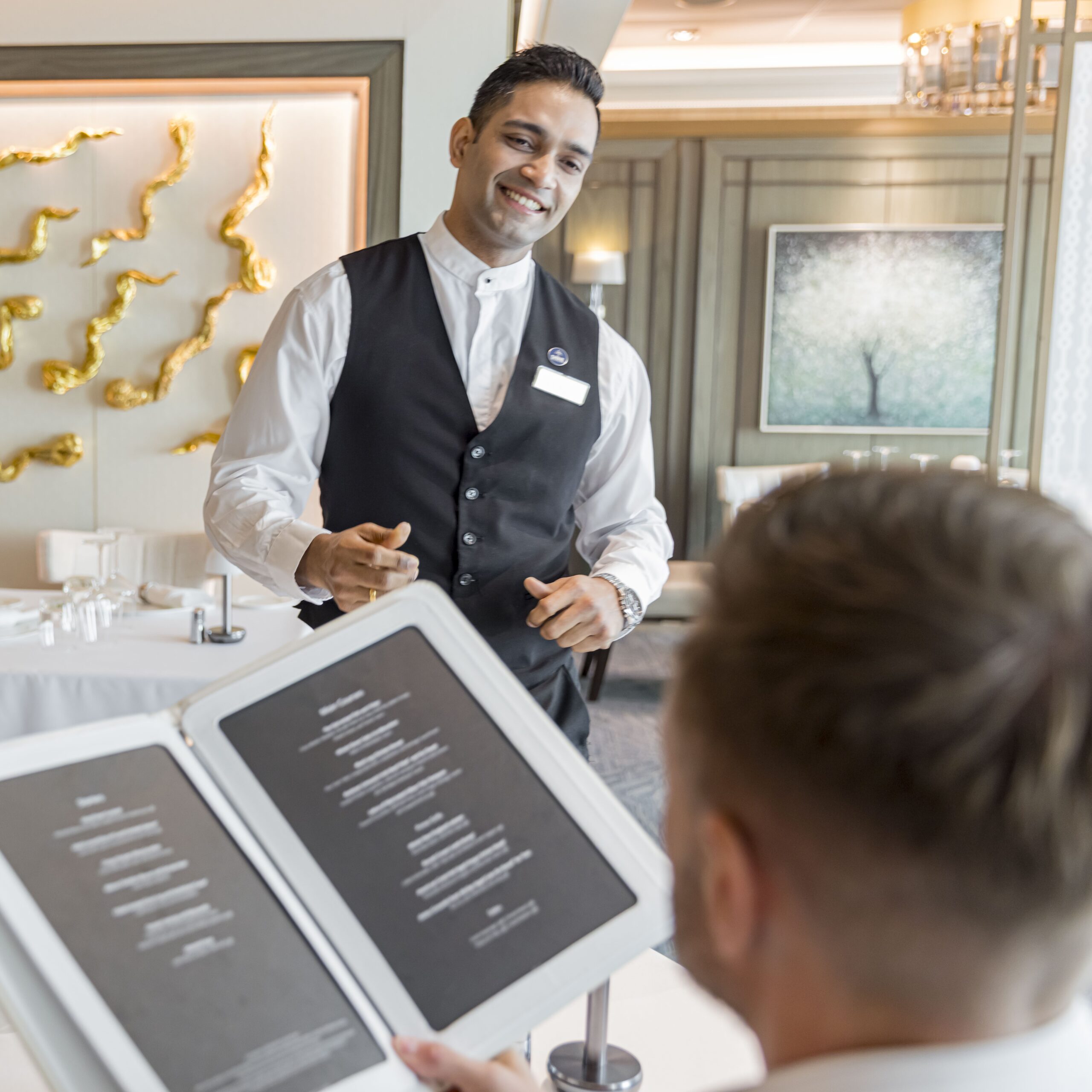 A smiling waiter in a black waistcoat stands beside a table, engaging with a guest holding an open menu in a modern, elegant setting—perfect for those interested in cruise ship restaurant jobs.