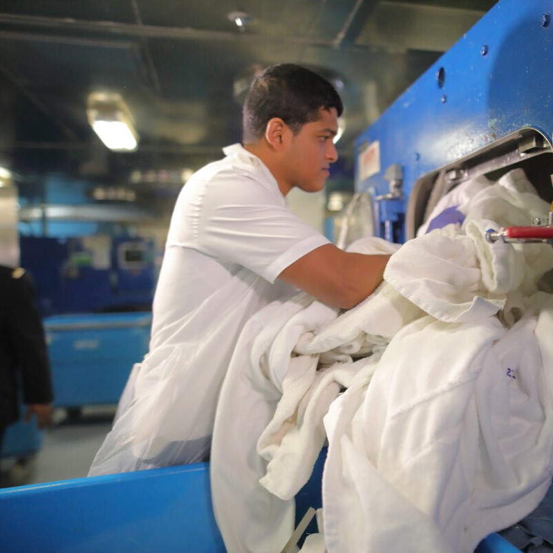 A man in a white uniform loads white towels into a large industrial washing machine in a laundry facility, surrounded by blue trolleys and equipment.