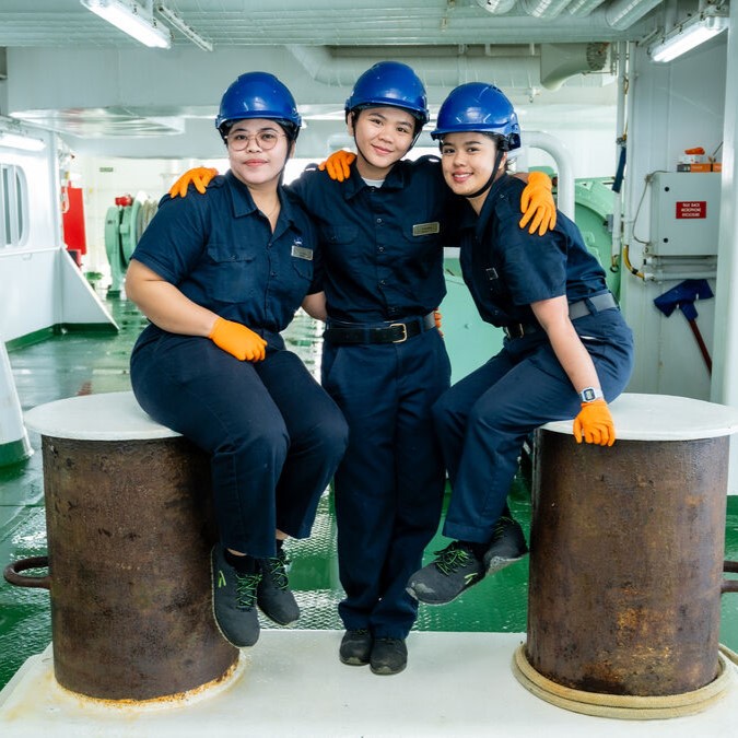 Three women in navy uniforms, blue helmets, and orange gloves pose together on a ship. Two are seated on large metal bollards whilst one stands between them, smiling with arms round each other—proudly representing P&O Cruises deck careers.