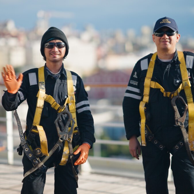 Two workers wearing black uniforms, yellow safety harnesses, and gloves stand outdoors on a rooftop—reminiscent of P&O Cruises deck careers. One waves and smiles; both wear sunglasses and hats against a blurred cityscape background.