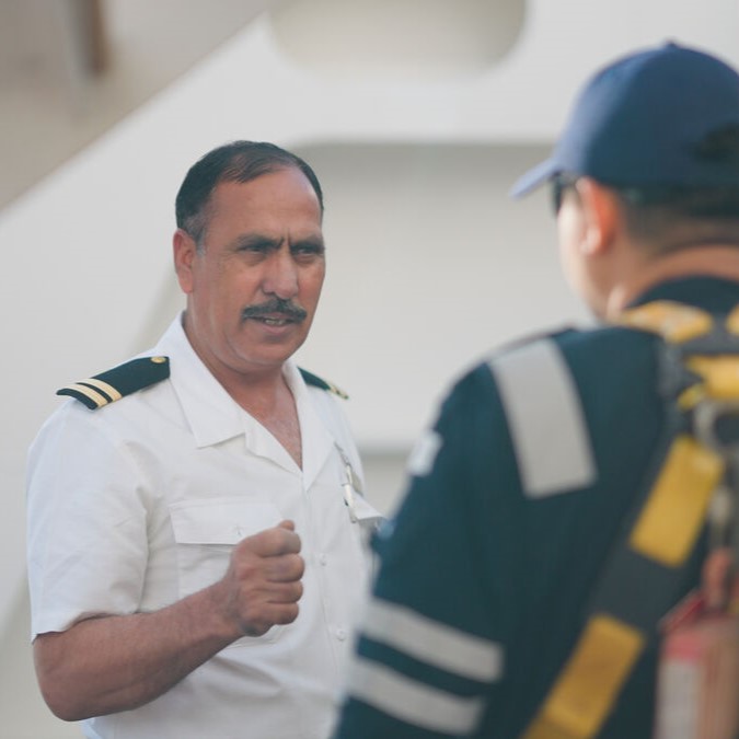 A man in a white uniform with shoulder stripes speaks to another person in a dark uniform and cap, seen from behind. The outdoor interaction suggests a glimpse into P&O Cruises deck careers on board a ship.