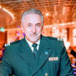 A man with grey hair and a beard, dressed in a dark green double-breasted uniform with captain's stripes on the shoulders, stands indoors with warm lighting and a blurred background.