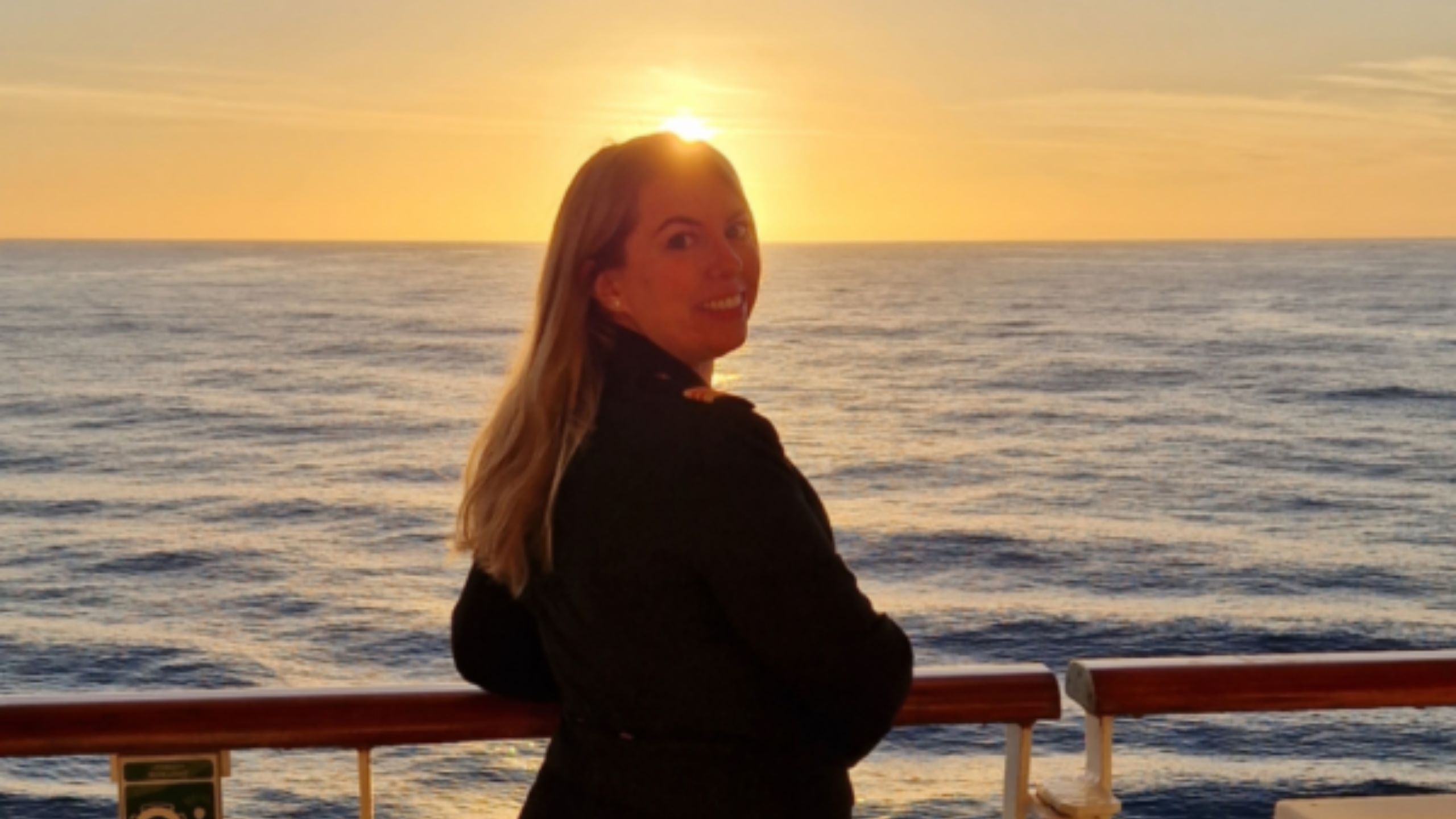 A ship's nurse stands on a ship’s deck at sunset, looking towards the camera. The golden light sets a tranquil scene—perfect for those interested in Cruise Ship Nurse Jobs and life at sea. A life buoy and railing are visible.