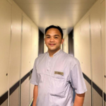 A person wearing a light grey uniform stands and smiles in a well-lit corridor with cream-coloured walls and doors on both sides.
