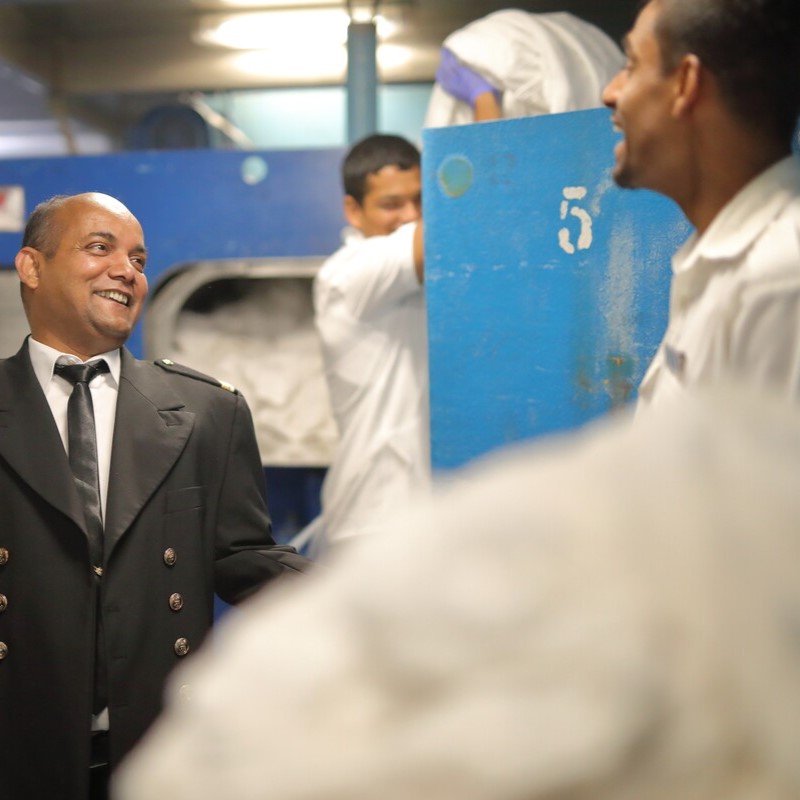 A man in a dark uniform with gold buttons smiles and talks to another man in white work clothes in an industrial laundry room, with another worker handling laundry in the background.
